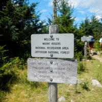 A photo of a sign at the trailhead. In the background are teenagers hiking into the woods.
