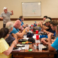A group of people eat lunch while listening to a man give a lecture in front of a whiteboard.