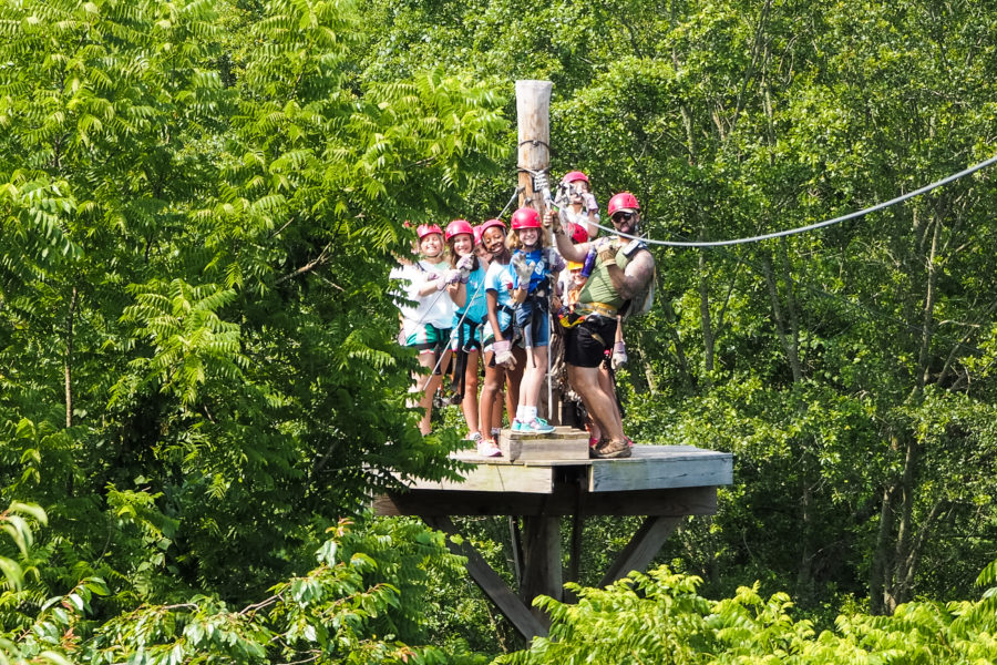 A group of resident campers stand on a tree top platform getting ready to zip line.