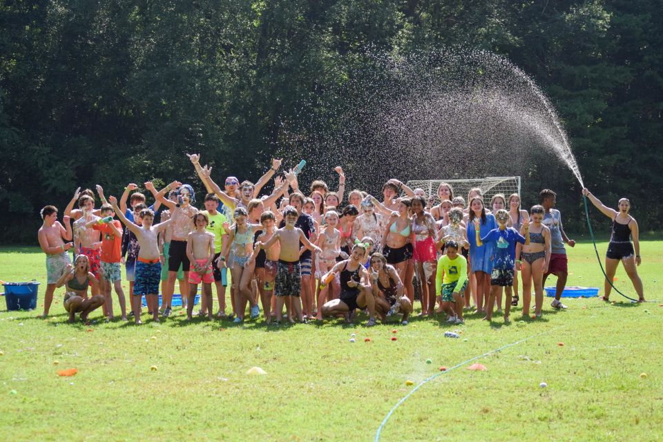 counselor holding hose sprays water over group of campers on the sports fields at Camp Canaan