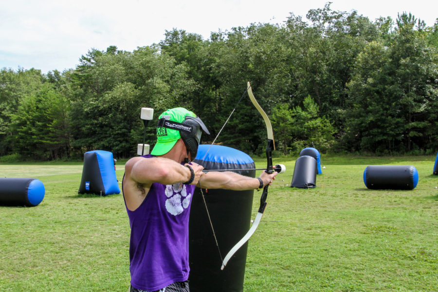 A teenager in a purple tank top takes aim with his bow and arrow during a game of archery tag.