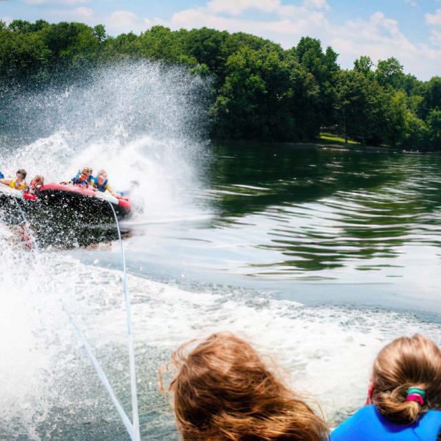 A group of people riding an inflatable tube on a lake, being pulled by a boat during summer camp. The boat's wake creates a large splash, adding excitement to the scene. The surrounding background features lush, green trees under a bright, sunny sky.