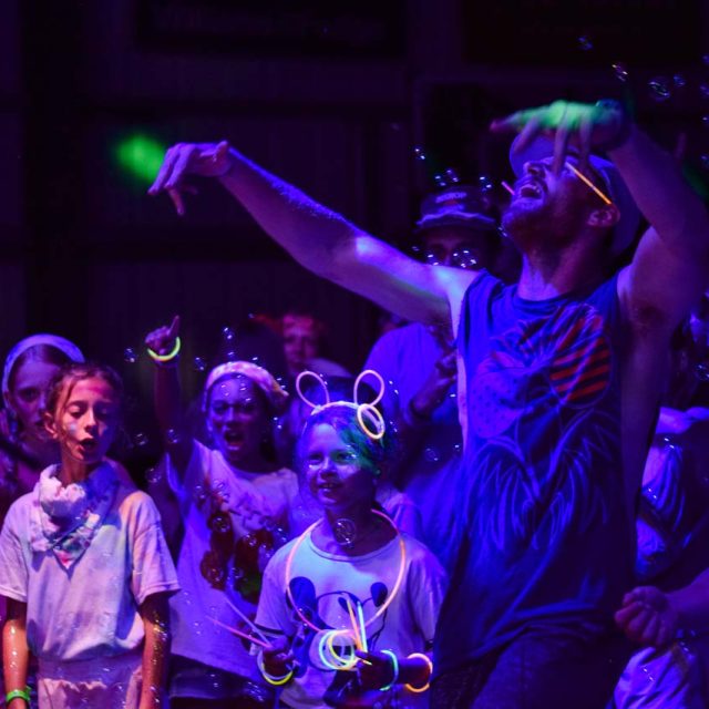 A lively group of campers and counselors at Camp Canaan glow under blacklight in the dimly lit covered basketball court. Their clothing and accessories fluoresce brightly. Some individuals have bubbles and glow sticks, while a man in a tank top energetically raises his arms, capturing the essence of summer camp fun.