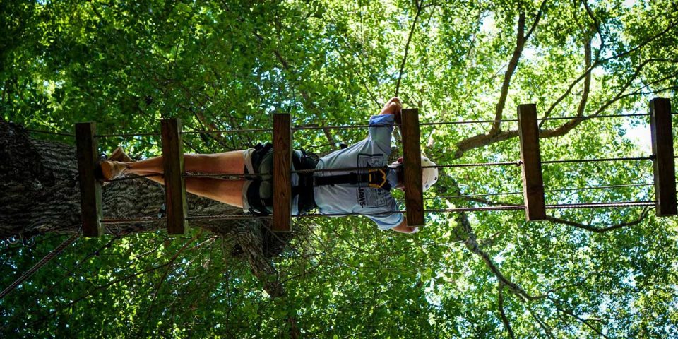 A person wearing a helmet and harness lays across a wooden plank bridge on the high ropes course.