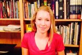 young blonde woman in red shirt stands in front of bookshelf