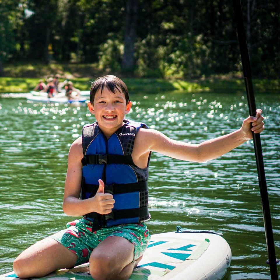 male senior day camper gives thumbs up on stand up paddle board floating on Lake Canaan