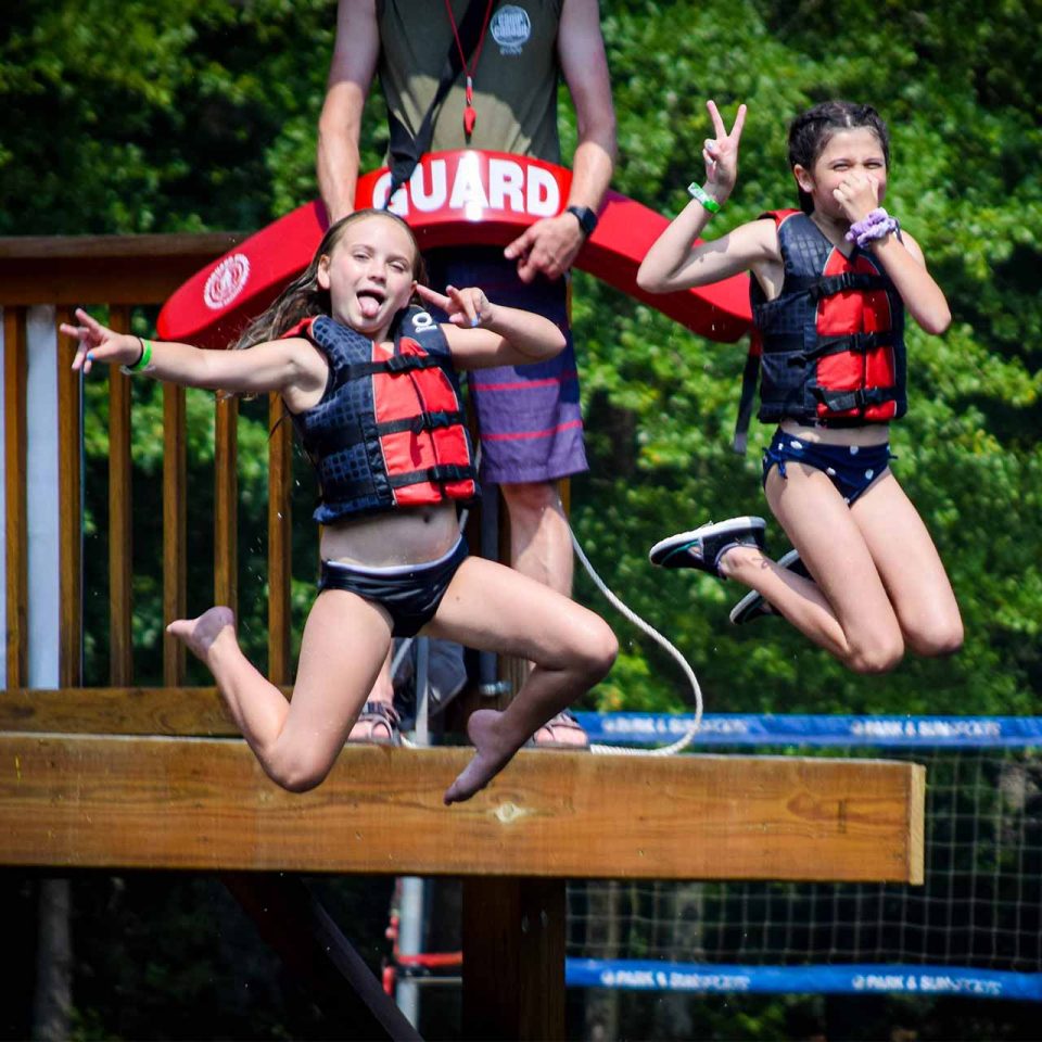 young girl campers pose mid-jump after jumping off of double-decker dock at Lake Canaan