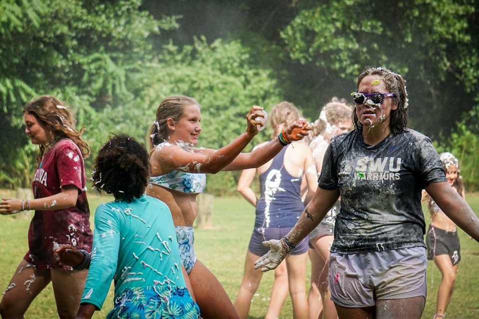 female counselor in purple sunglasses walks through groups of campers covered in shaving cream after sludge war