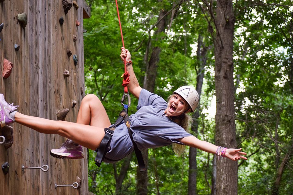 young woman in white helmet leans back from climbing wall while on belay in her harness