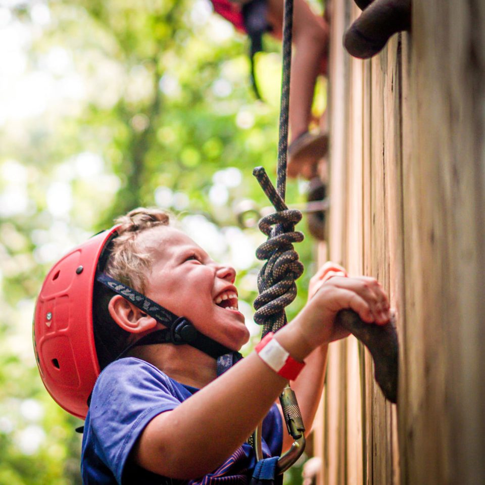 junior day camper wearing red helmet smiles while rock climbing at Camp Canaan