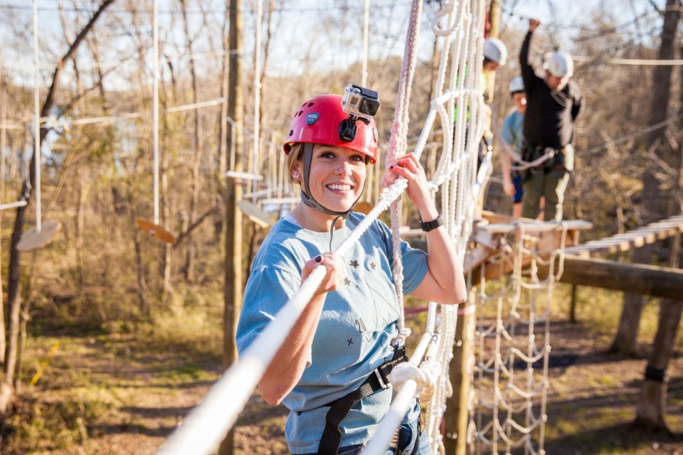 young woman with GoPro installed on red helmet walks across high ropes course element at Camp Canaan