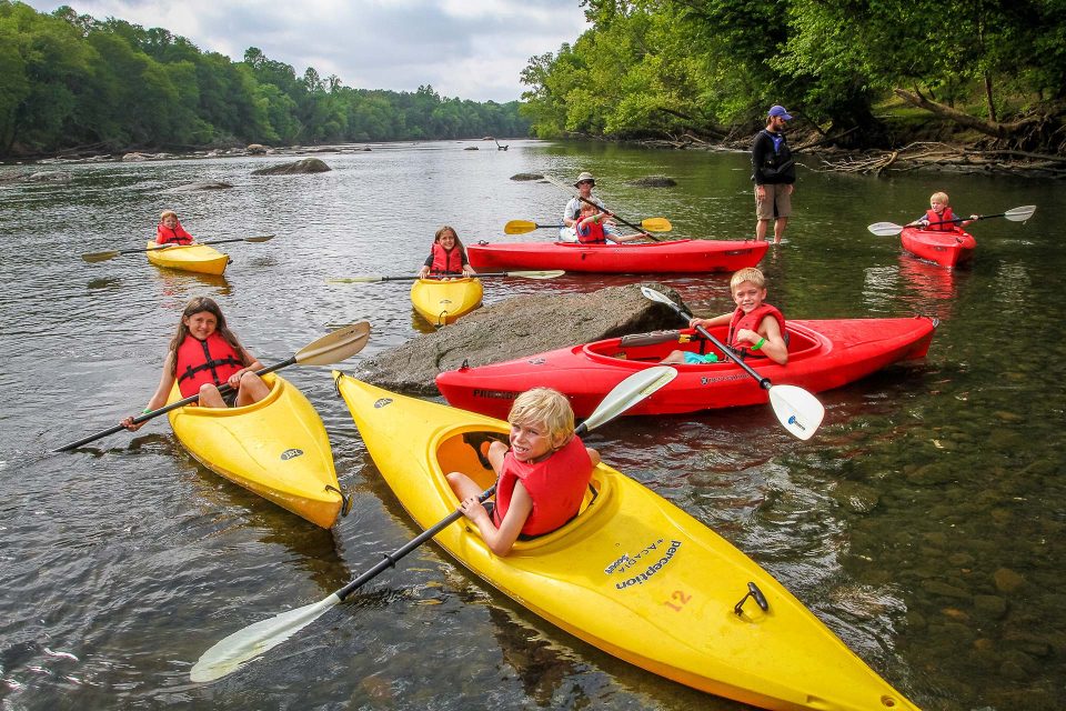 seven smiling kids in red and yellow kayaks hold their paddles to smile at the camera