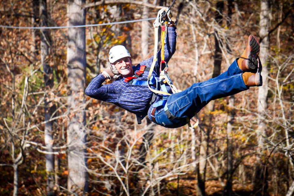 man in white helmet poses while zipping past fall-colored leaves on the Zipline Canopy Tour at Camp Canaan