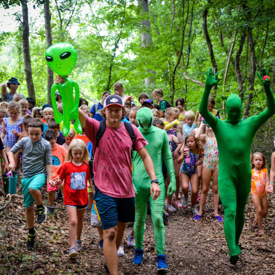 counselor holding inflatable green alien is joined by other counselors dressed as aliens leading junior day campers on a nature walk around Camp Canaan