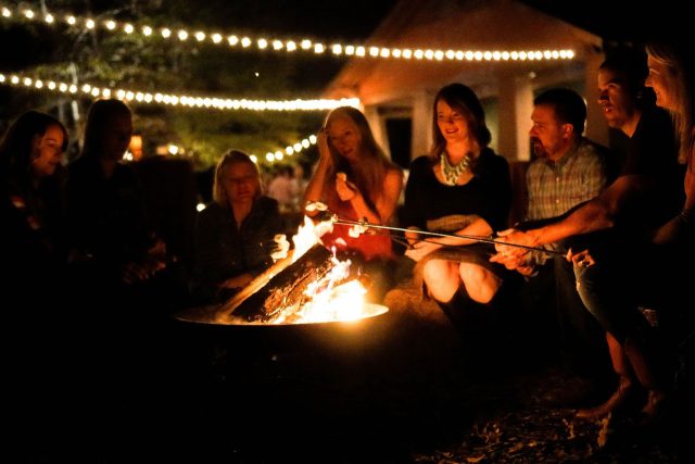 adults roast marshmallows over a bonfire under cafe bulb lighting outside of the multi-purpose building at Camp Canaan