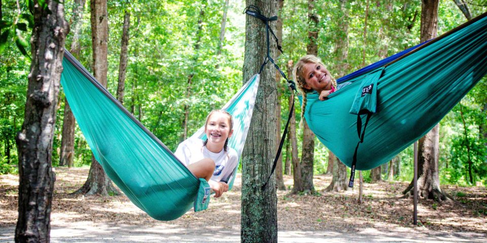 Two people are smiling and relaxing in teal hammocks that are hanging from trees in a wooded area at Camp Canaan. The ground is covered with leaves and dried foliage, while the surrounding green trees create a peaceful outdoor atmosphere, perfect for a summer camp.