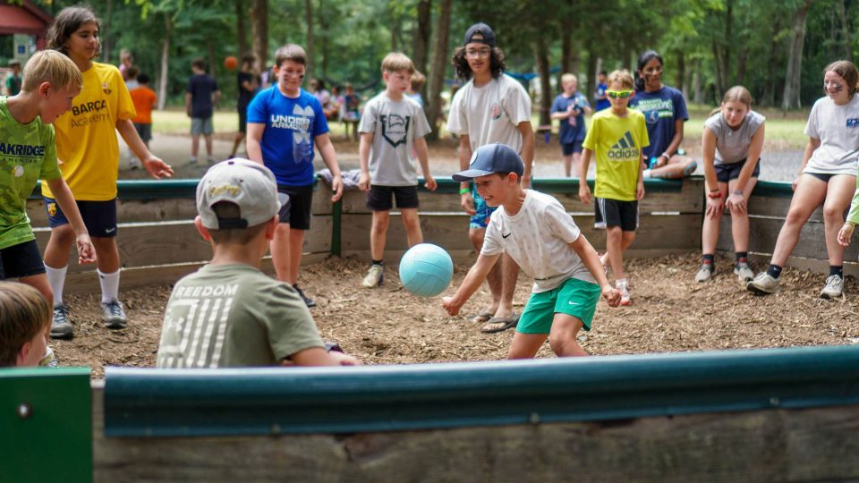 Children are actively playing a game of GaGa ball in an outdoor pit surrounded by wooden walls at the summer camp. Many children are waiting their turn, standing around the perimeter and watching the game. The scene is lively, with trees in the background.