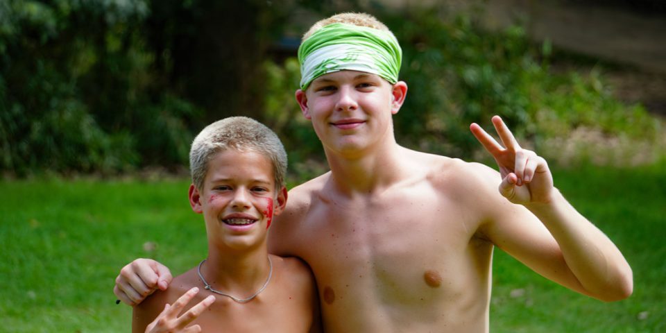 Two shirtless boys outdoors smile at the camera. The younger boy, on the left, with short, light-colored hair and braces, has his arm around the older boy, who wears a green bandana. Both boys hold up peace signs with their right hands. Greenery is visible in the background – it's pure Camp Canaan magic!