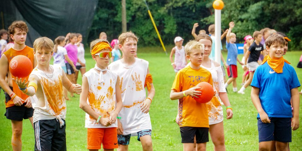 A group of children stands on the sports field at the summer camp, getting ready to play a game. They hold orange balls and wear colorful clothes, mainly in blue and orange. Some kids wear orange bandanas around their necks or on their heads. Trees and other campers are in the background.