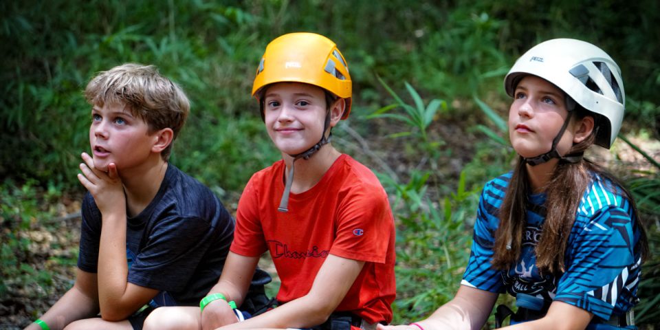 Three children sit outdoors at Camp Canaan, wearing helmets and casual clothing. They appear to be in a forest or wooded area. One child on the left is looking upwards with a thoughtful expression, the middle child is smiling, and the child on the right is also looking up, excited for their high ropes course adventure.