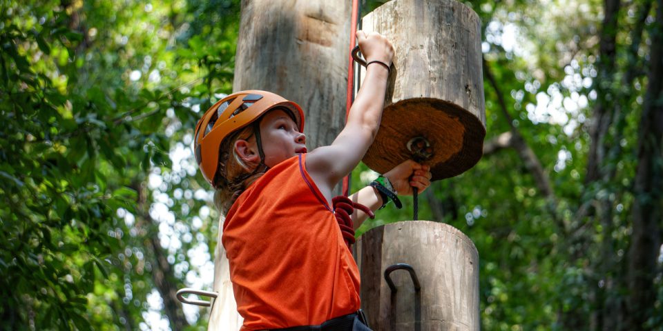 At Camp Canaan, a young girl wearing an orange helmet and shirt grips onto a large wooden post element on the Catawba Tower while climbing in the forest during summer camp. She is secured with a safety harness, and trees with green foliage surround her on a bright, sunny day.