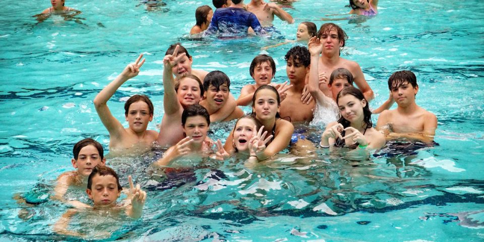 A group of children and teenagers in swimwear enjoying themselves in a pool at Camp Canaan. Many are smiling, making peace signs, and posing for the camera. The water is clear and blue, with more people visible in the background.