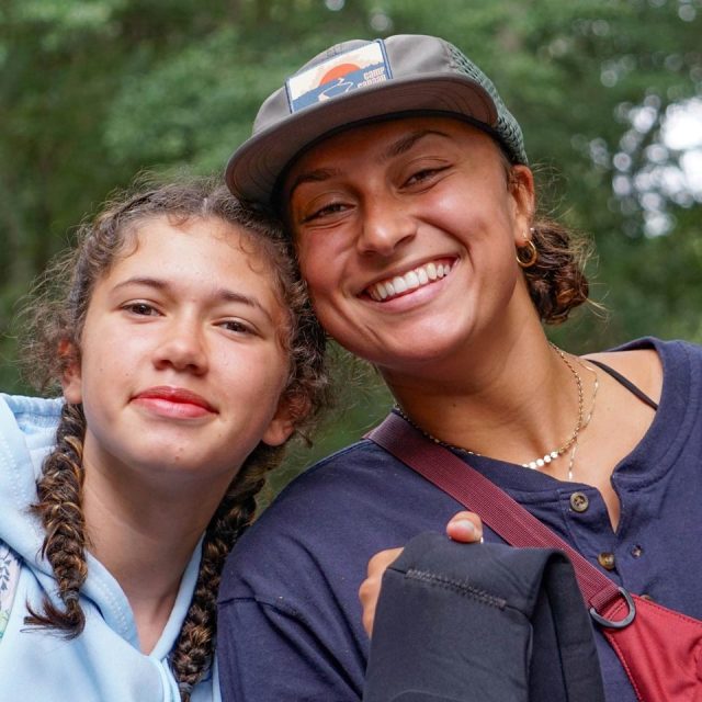 Two people are smiling and standing close together outdoors at an overnight summer camp. The person on the left has braided hair and is wearing a light blue hoodie with a colorful design. The person on the right is wearing a cap and carrying a bag and a beverage can. Trees are visible in the background.