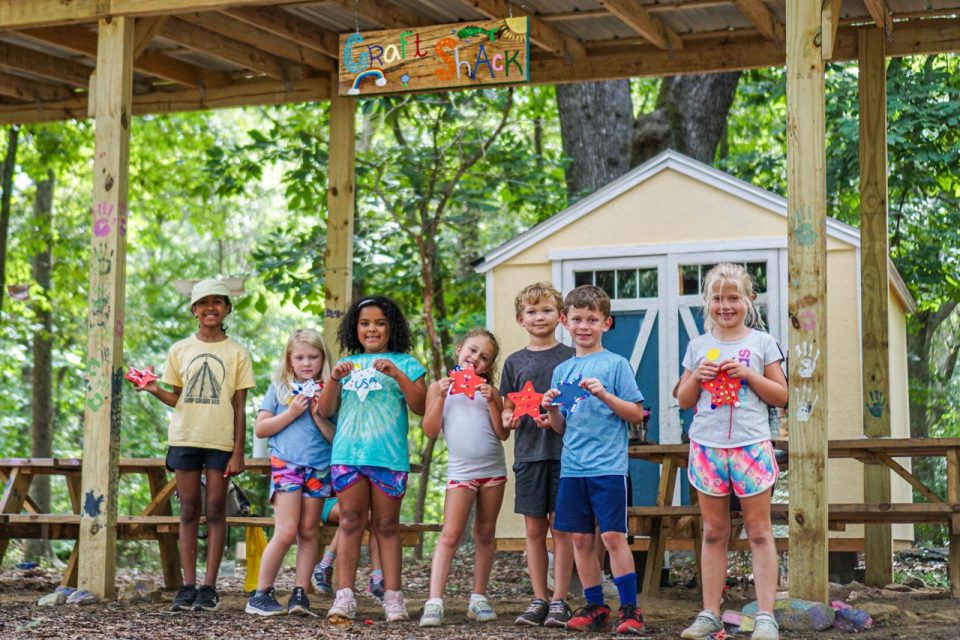 group of kids smile holding their crafts in front of the Craft Shack at Camp Canaan