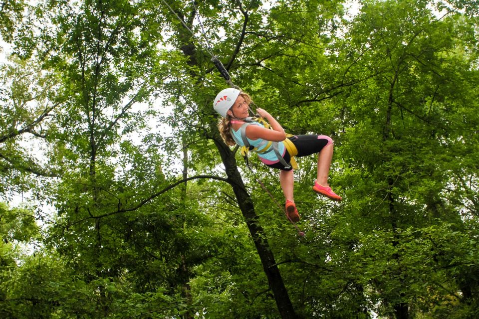 young female camper in white helmet and red shoes swings down from a high ropes course element at Camp Canaan