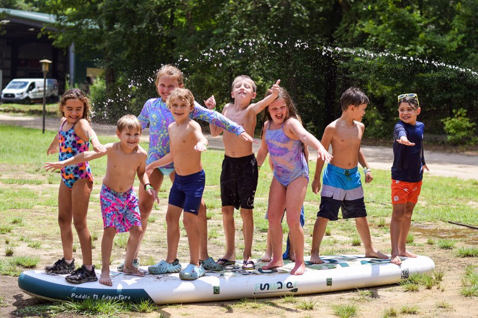 group of junior day campers pretend to surf on stand up paddle board laying on land next to Lake Canaan