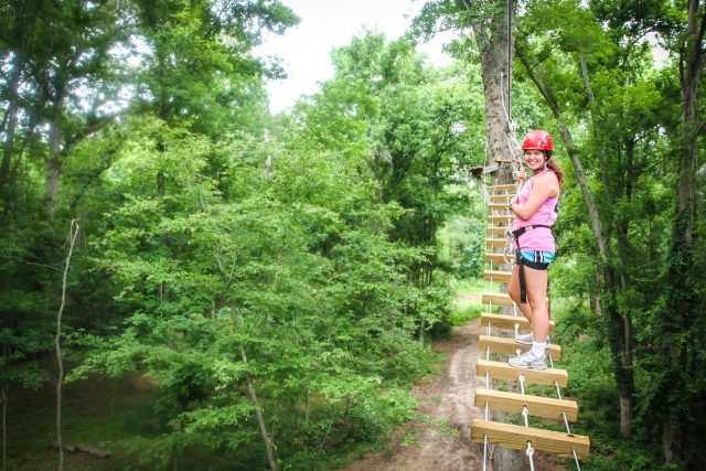 young woman in pink shirt and red helmet smiles back at camera while walking on a high ropes course element at Camp Canaan