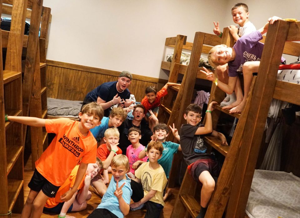 A group of children and two counselors pose playfully for a photo in Camp Canaan's A&J lodge cabin during summer camp. The kids are smiling, making peace signs, and some are leaning out of the bunks.