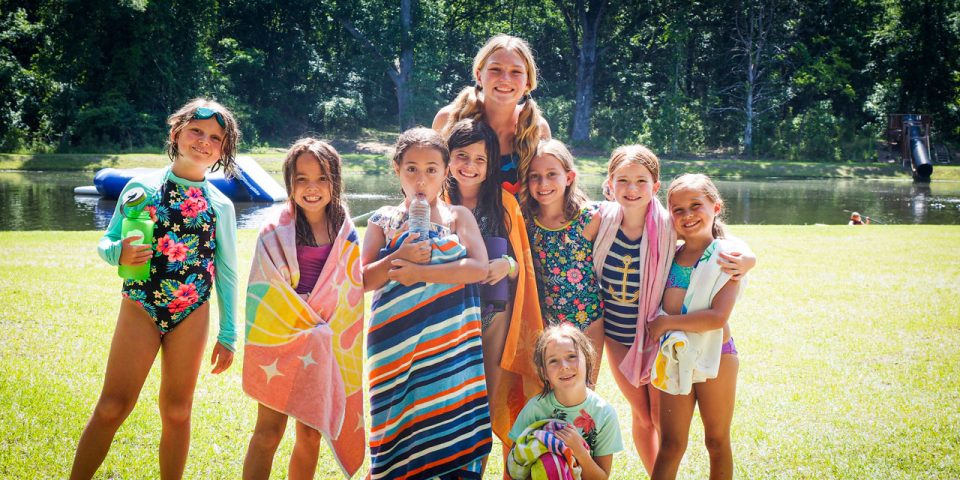 A group of smiling girls in swimsuits stand on grass near the lake at Camp Canaan. Some are wrapped in towels. A tall female counselor stands in the middle behind the children. Trees and the lake are visible in the background. It's a sunny day at this vibrant summer camp.