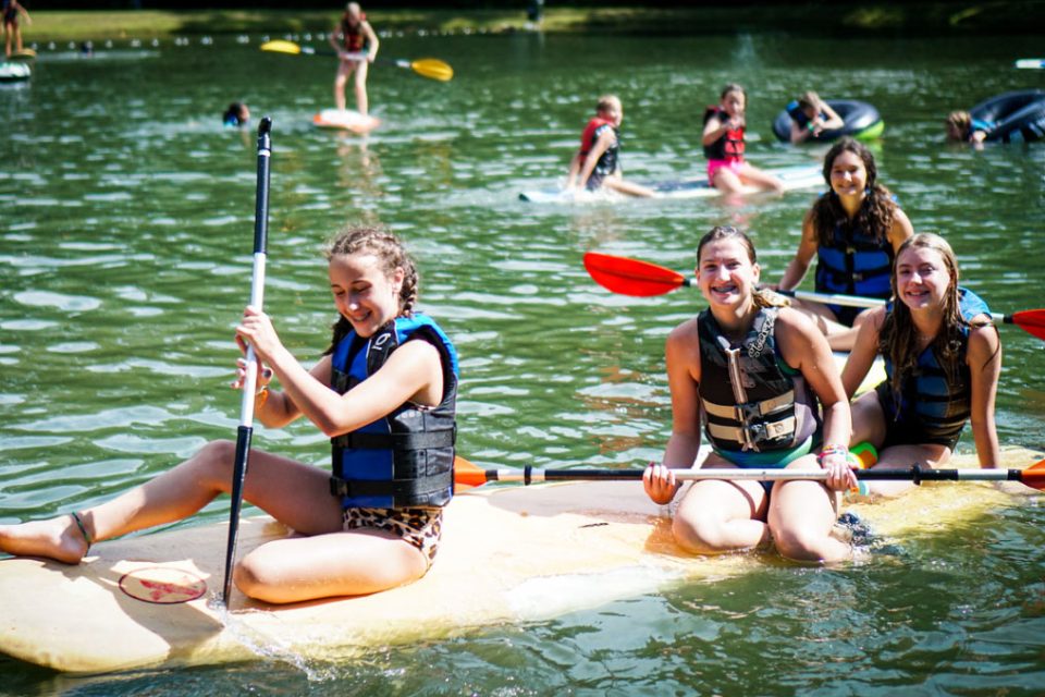 young female campers sit on stand up paddle board together in Lake Canaan holding paddles