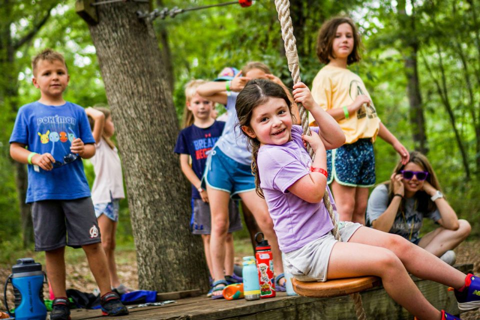 junior day camper in purple shirt smiles as she rides down the zipline on the playground at Camp Canaan