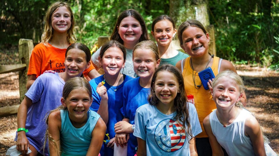 A group of ten smiling girls pose together outdoors at Camp Canaan, with trees and a wooden fence in the background. They're wearing casual summer clothing for summer camp, and several girls are sporting playful expressions.