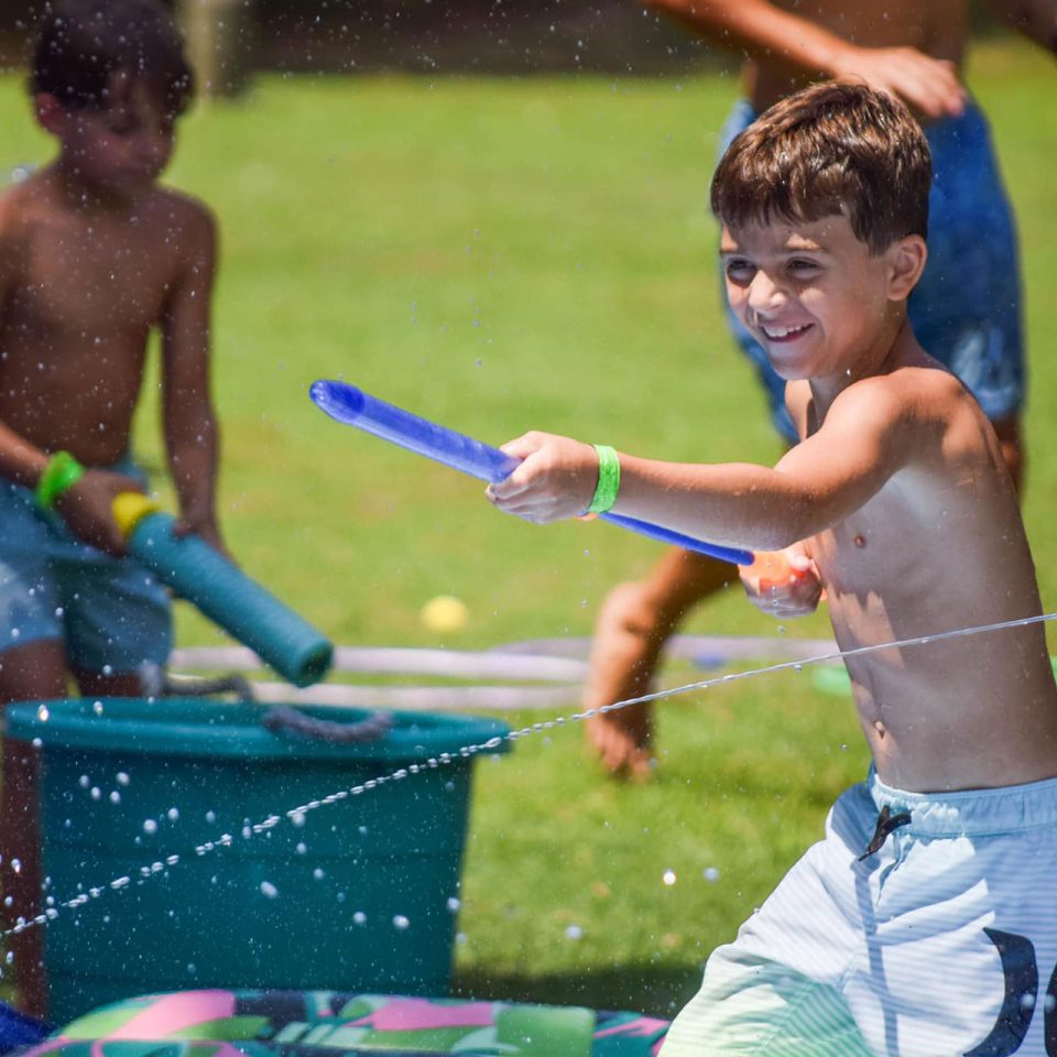 young male camper squirts water from a blue water toy on the Camp Canaan sports field