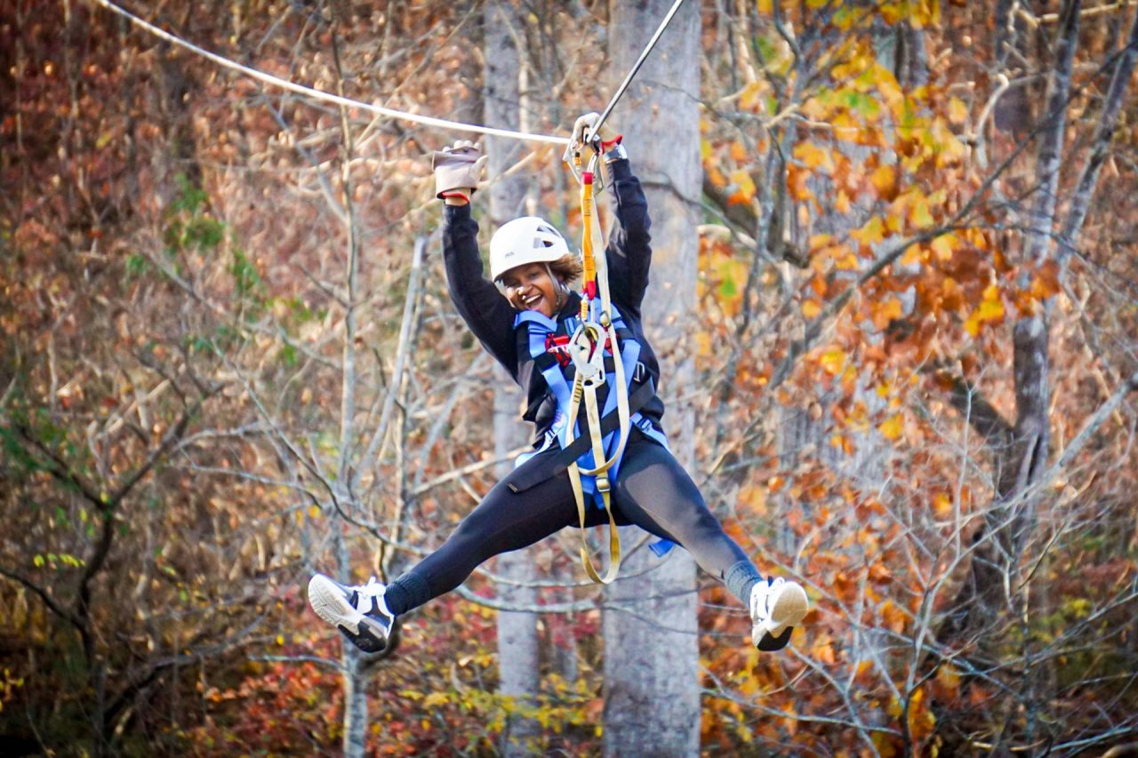 young woman in white helmet smiles and does a split while zipping on the Zipline Canopy Tour at Camp Canaan