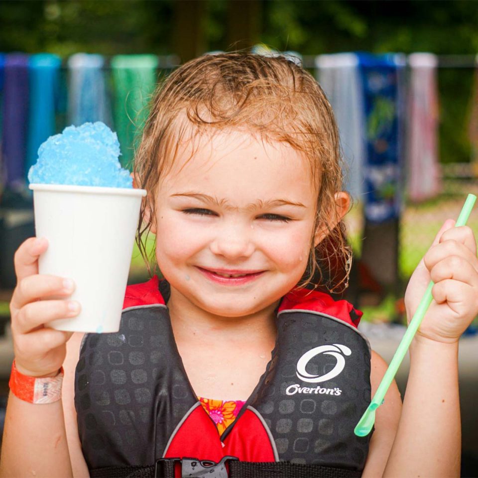 junior day camper holds up blue snow cone and straw wearing life vest during summer camp at Camp Canaan