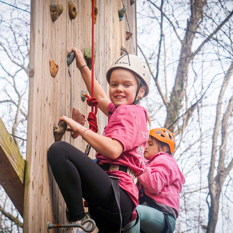 female camper in pink shirt and white helmet climbs up the rock climbing wall during winter camp at Camp Canaan