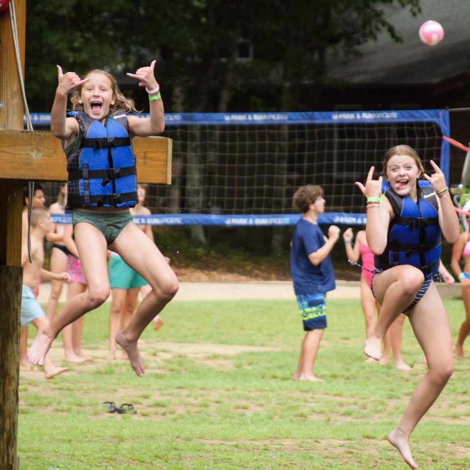 overnight girl campers pose for the camera mid-air on the volleyball court by Lake Canaan