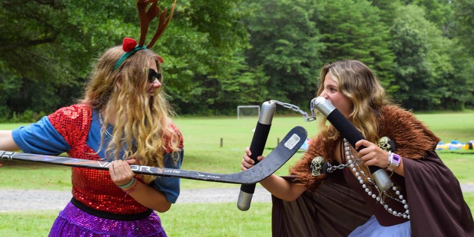 Two people dressed in whimsical costumes playfully pretend to duel with a hockey stick and nunchucks at Camp Canaan's summer camp.