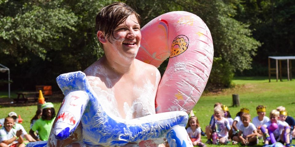 A young man with a joyful expression stands outdoors at Camp Canaan, covered in foam and wearing an inflatable ring. In the background, a group of children sits on the grass, some wearing playful hats or foam. Trees and greenery surround the lively summer camp scene.