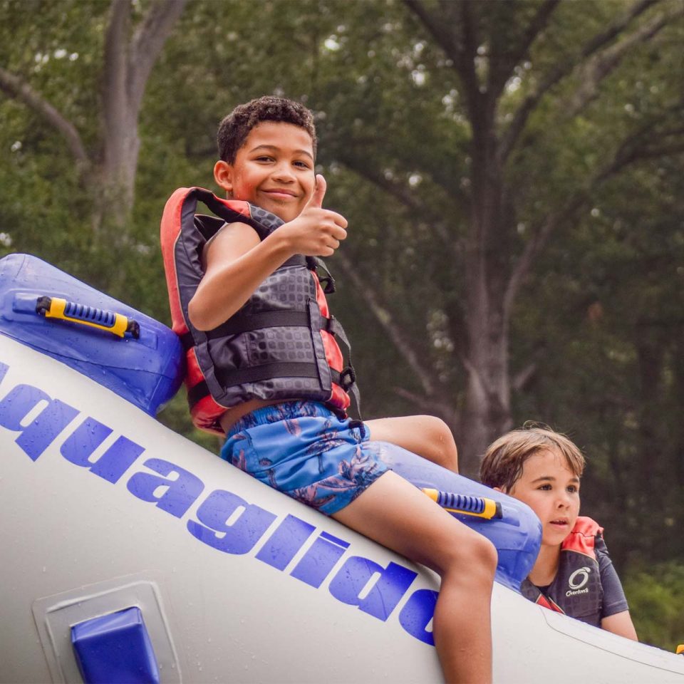 male junior day camper gives thumbs up on top of inflatable raft in the lake at Camp Canaan