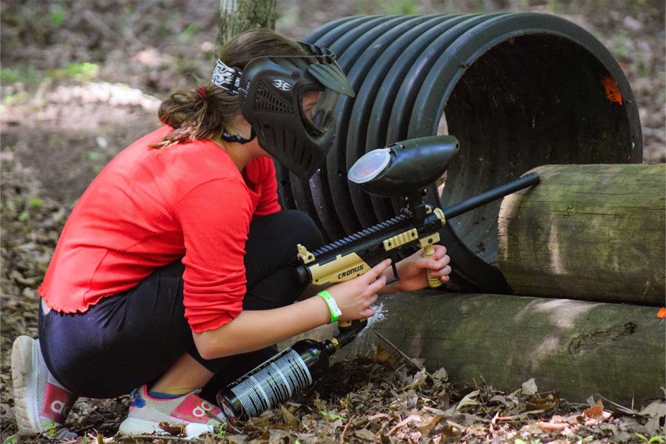 girl in red shirt crouches behind logs preparing for her next paint ball shot