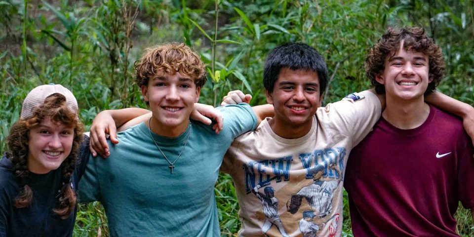 Four young people stand closely together outdoors, smiling at the camera during summer camp. With arms around each other's shoulders, they look happy and relaxed. The background features green foliage.