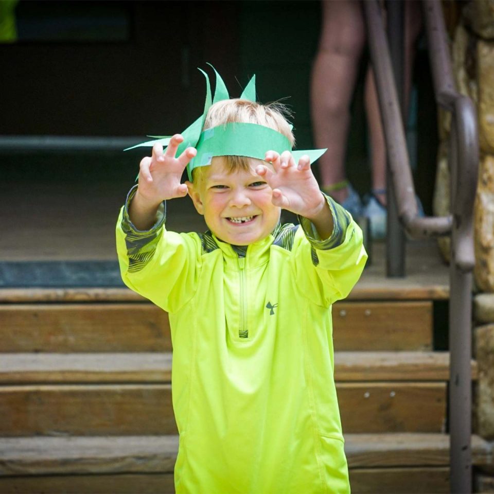 junior day camper makes claws with hands while wearing green handmade dino hat