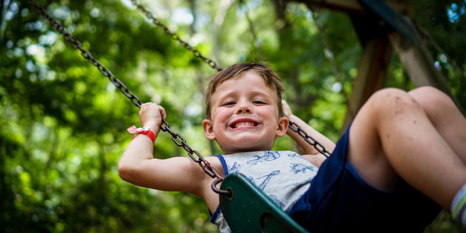 junior day camper smiles while playing on the swings at the playground at Camp Canaan