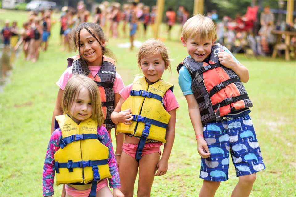 group of smiling junior day campers wearing bright life vests by the lake at Camp Canaan