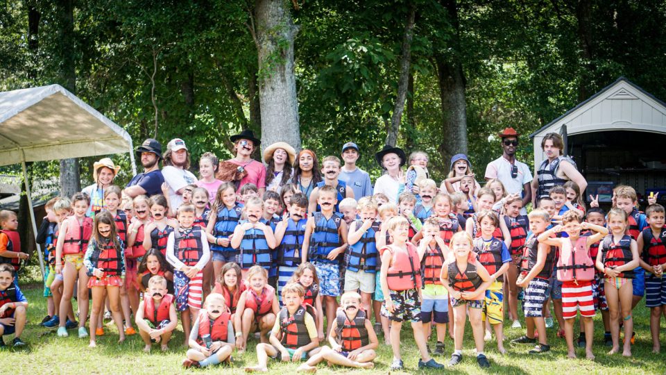 large group of campers and counselors wear life vests and fake moustaches by the lake at Camp Canaan